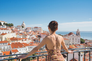 A tourist woman looks at the beautiful cityscape of Lisbon, with the colorful houses and roofs at the Alfama district, Portugal. - obrazy, fototapety, plakaty