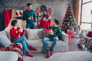 Full body portrait of group peaceful friendly people sitting couch drink cacao hold new year giftbox communicate decorated tree indoors