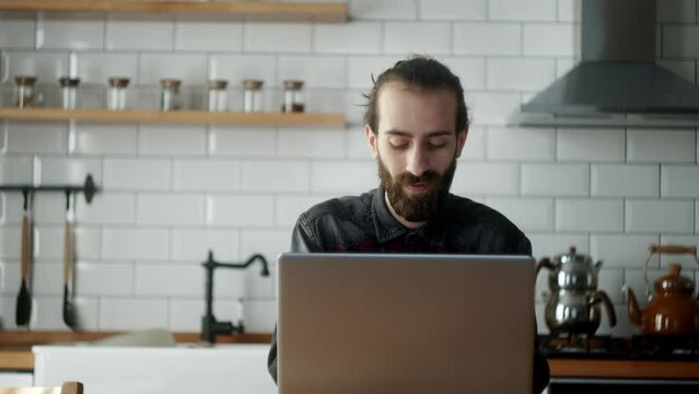Bearded young adult freelancer man sitting in the kitchen working with laptop, getting works done