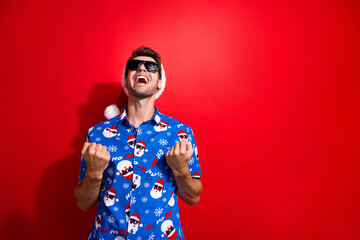 Portraitof satisfiec man dressed christmas hat blue shirt in sunglass clenching fists win lottery isolated on vivid red color background