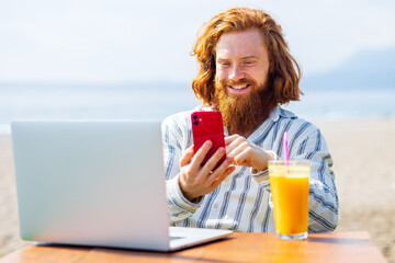 young redhaired ginger bearded man working outdoors in sea cafe with laptop an summer day