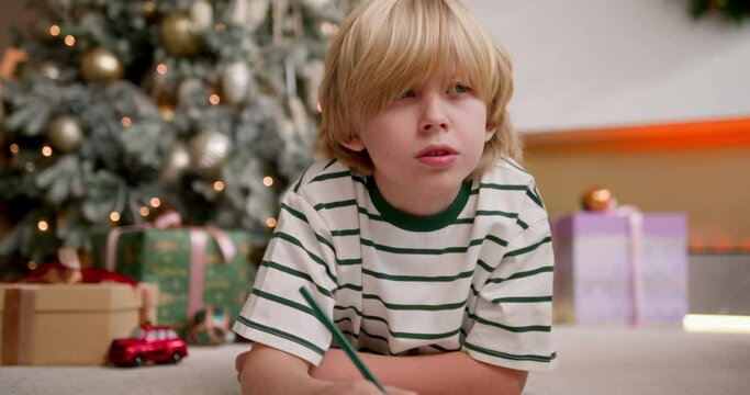 blonde boy writes a letter with wish list to santa claus using color pencils laying on floor near christmas tree.