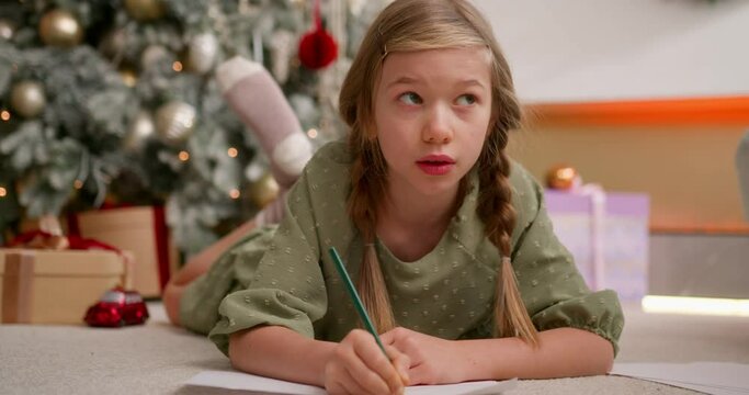 girl writes a letter with wish list to santa claus using color pencils laying on floor near christmas tree.