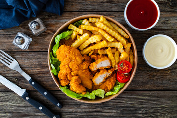 Fried breaded chicken nuggets served with French fries and vegetables on wooden table
