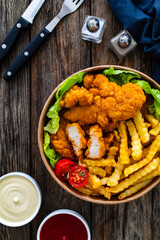 Fried breaded chicken nuggets served with French fries and vegetables on wooden table

