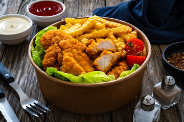 Fried breaded chicken nuggets served with French fries and vegetables on wooden table

