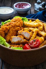 Fried breaded chicken nuggets served with French fries and vegetables on wooden table
