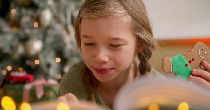 girl lays on floor near christmas tree, reads book of magic stories and bites gingerbread man cookie. Turns over pages chewing and smiling.