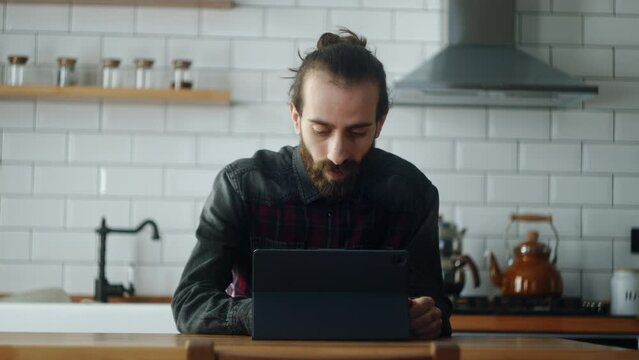 Bearded young adult man sitting in kitchen, using app with tablet device, surfing on internet, spending free time