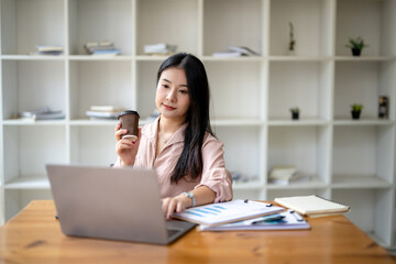 Fototapeta na wymiar Asian business woman sitting and working in front of a laptop, holding a cup of coffee work independently and relax.