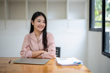 Asian businesswoman sits with her arms crossed at a desk with documents on the table.