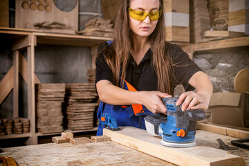 Young woman builder carpenter equals polishes wooden board with a  random orbit sander  in the...