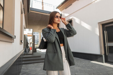 Fototapeta na wymiar Beautiful fashion business girl in fashionable clothes with a stylish green blazer with a bag puts on glasses and stands in the city near the building
