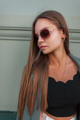 Naklejka premium Street female portrait of a beautiful fashionable young business woman model with vintage sunglasses in fashion elegant clothes stands near a green wall outdoors