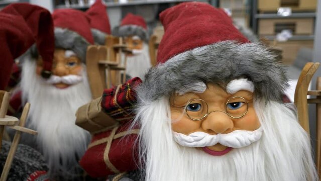 Many Santa Claus dolls standing in rows, in the store interior during the Christmas sale. Panning shot of white-bearded smiling Santas with red gift boxes, and wooden skis, on the shopping mall shelf.