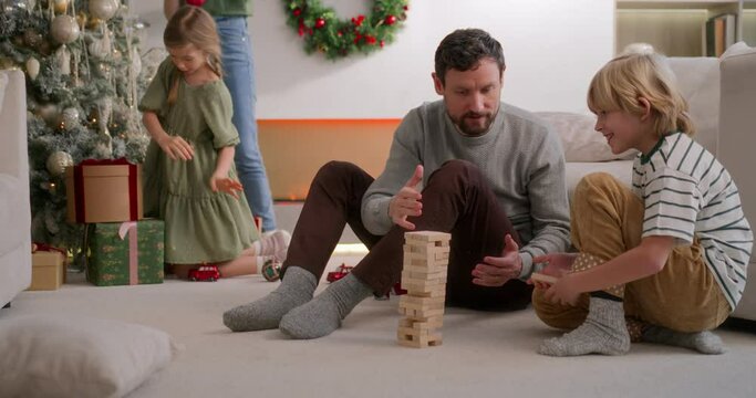 dad and son play jenga together on floor at home, smiling and laughing, mom and daughter decorate christmas tree on background