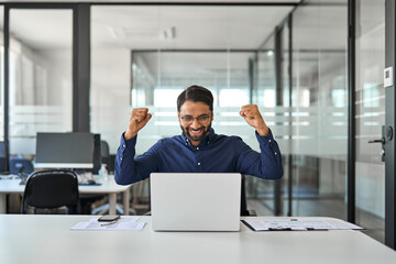 Happy professional Indian businessman employee raising fists celebrating win using laptop. Excited...