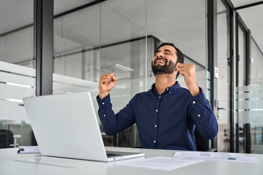 Happy excited professional Indian business man employee executive celebrating win online and financial success screaming yes using laptop computer feeling motivated at work in office.