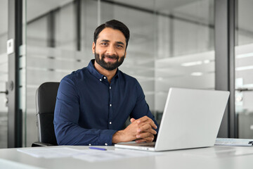 Smiling confident Indian business man employee looking at camera sitting at work desk with laptop computer. Portrait of smart happy businessman office worker or entrepreneur posing at modern workplace