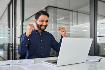 Happy Indian business man employee manager winning prize online working using laptop in office. Excited businessman executive looking at computer celebrating success, project growth, getting profit.