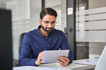 Busy Indian business man manager using tablet computer, businessman employee or executive looking at tab device analyzing finance market data working with ai solutions sitting at office desk.