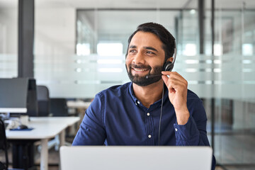 Smiling happy Indian call center agent wearing headset talking to client, contract service telemarketing operator using laptop having conversation working in customer tech assistance support office.