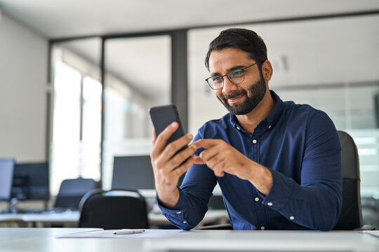 Busy Indian business man executive using mobile phone working in office. Professional businessman employee looking at smartphone checking banking financial applications sitting at workplace desk.