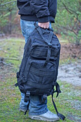 the hand of a man in blue jeans holds a black army backpack in his hand on the street