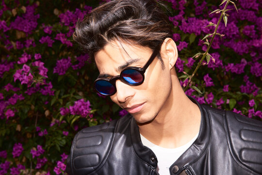 Portrait of a handsome smiling Latin man wearing sunglasses and leather jacket looking down posing in front of a background of flowers