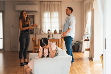 Annoyed and unhappy young girl sitting on sofa trapped in middle of tension by her parent argument...