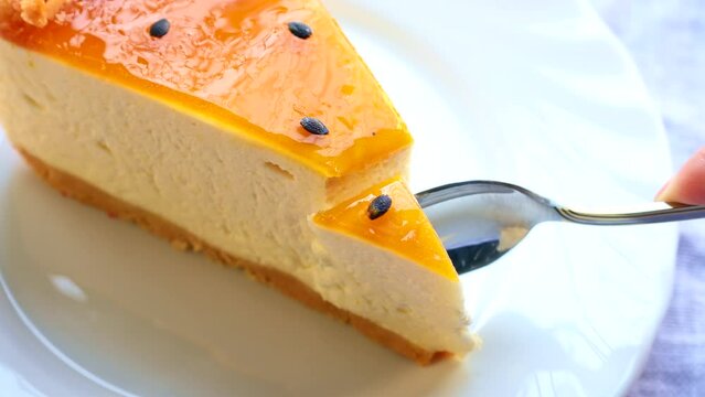 Eating delicious fresh baked multi-layered mango passionfruit cheesecake slice with fruit jelly glaze on white plate with a spoon close up