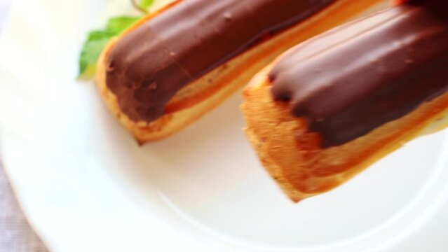Hand taking French pastry eclairs with cream filling covered with melted milk chocolate and mint leaves from a white plate close up
