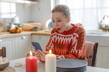 young blonde woman sits at table in kitchen with Christmas decorations, gets massage with unexpected great news, feels elation and thrilled. unbelievable and astonishing information