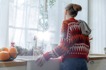 young woman in red sweater stays in kitchen leaning on sink feeling upset. thoughtful and frustrated girl at home during homework looks through window at Christmas holidays