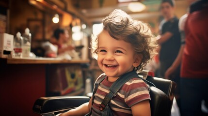 A 3-year-old boy wearing a protective cape sits in a happy barber's chair while getting a haircut in a cutely decorated shop for children. - obrazy, fototapety, plakaty