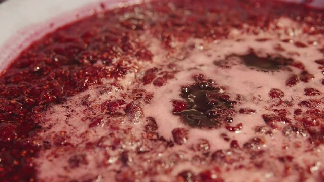 A detailed shot of raspberry jam in the process of simmering, showing glistening bubbles and a deep red hue, foreshadowing its transformation into a homemade preserve.