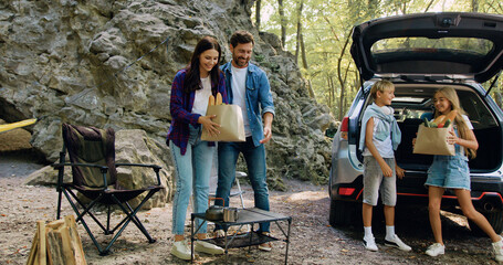 Parents and children take out unpacking packages of products from open trunk of car in forest. Concept of vacation, family travelling and bonding. Beautiful smiling family is preparing for picnic in - obrazy, fototapety, plakaty