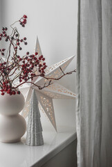 Scandinavian style Christmas window decor. Scandinavian paper star, ceramic Christmas tree, cranberry branches in a vase on the windowsill