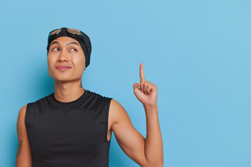 Young swimmer stands on blue background, pointing upwards, smiling, advertising space, sport life...