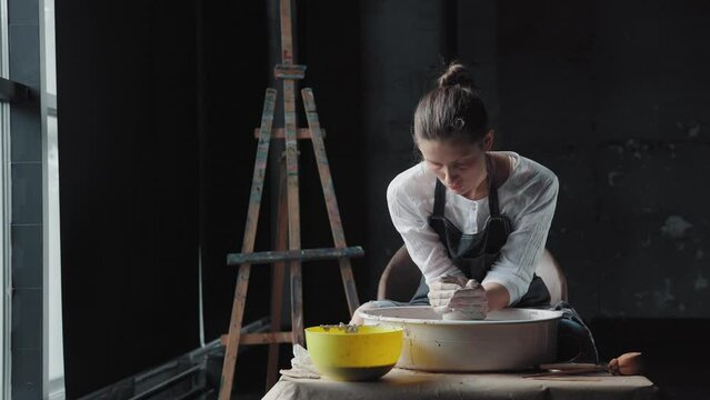 A woman potter in a black apron and shirt works in workshop with a potter's wheel, a general plan. Pottery workshop: young woman and potter's wheel. Easel on the background.