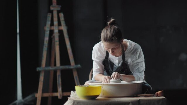 A woman potter in a black apron and shirt works in workshop with a potter's wheel, a general plan. Pottery workshop: young woman and potter's wheel. Easel on the background.