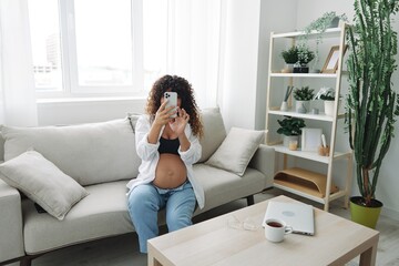 Pregnant woman blogger sits on the couch at home and takes pictures of herself on the phone, selfie and video call, online doctor consultation, pregnancy management, freelance work smiling