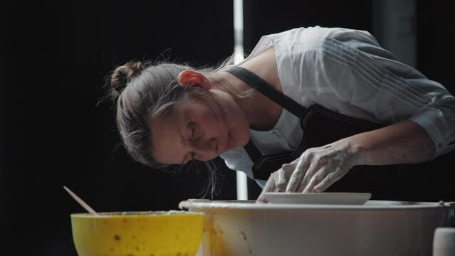 A young female potter in the workshop: she works with a potter's wheel, leaned over the product and carefully makes the product. Dark studio, natural light.