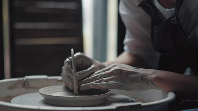 Woman potter makes a plate on a potter's wheel. Hands in white clay. Medium plan, shot from the side. Potter's workshop.