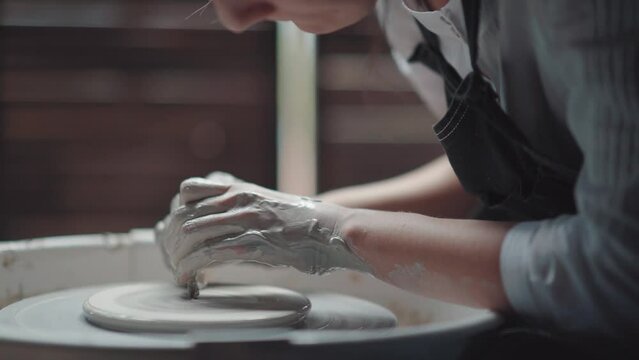 A woman potter in an apron bent over a potter's wheel. Her hands are in clay. Medium plan, shot in a creative workshop.