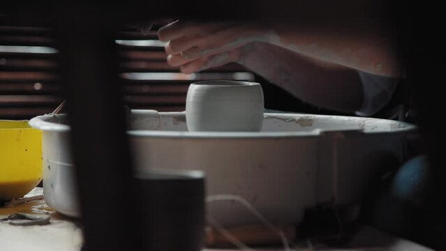 A woman potter in a black apron bent over a potter's wheel and makes a pot or mug. She works in a workshop with natural light. Medium shot.