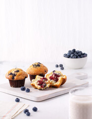 Blueberry streusel muffins on a white cutting board with a bowl of fresh blueberries in the background and a glass of milk in the foreground; copy space