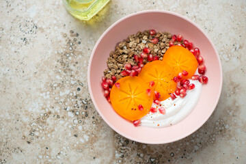 Roseate bowl with persimmon slices, granola, yogurt and pomegranate, horizontal shot on a beige granite background with space, top view