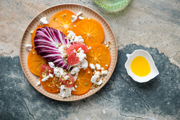 Fresh persimmon with radicchio, goat cheese, grapefruit and pistachios, flat lay on a grey and beige granite background, horizontal shot - obrazy, fototapety, plakaty
