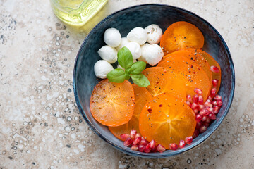 Blue bowl with fresh sliced persimmon, mini mozzarella and pomegranate, horizontal shot on a beige granite background, elevated view